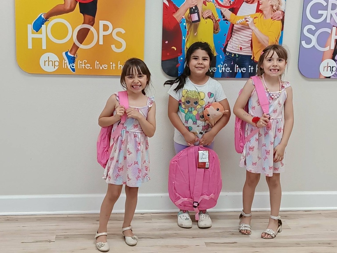 Three girls stand holding pink backpacks from the Bayshore backpack giveaway